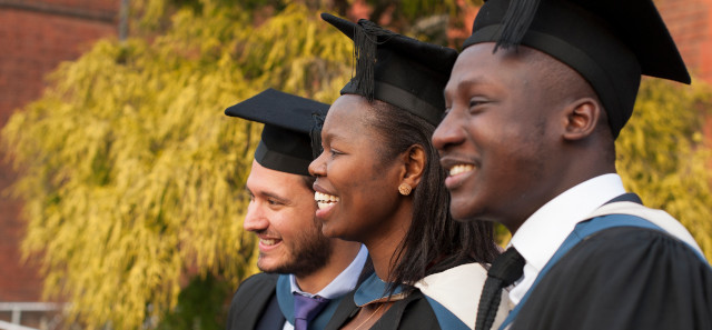 Three smiling students stand in a row wearing their graduation cap and gowns. 